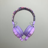 A pair of purple over-ear headphones with a colorful patterned headband, held in a person's hand against a tiled floor background.